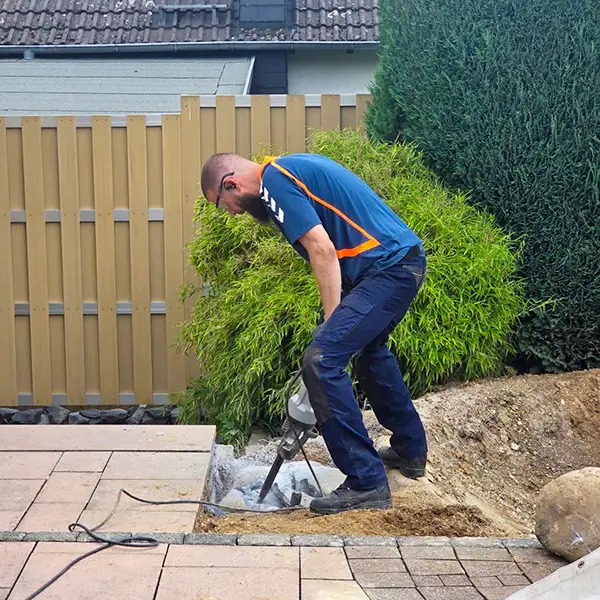 Ein Mann in blauer Bekleidung arbeitet mit einem elektrischen Meißel auf Steinplatten im Garten